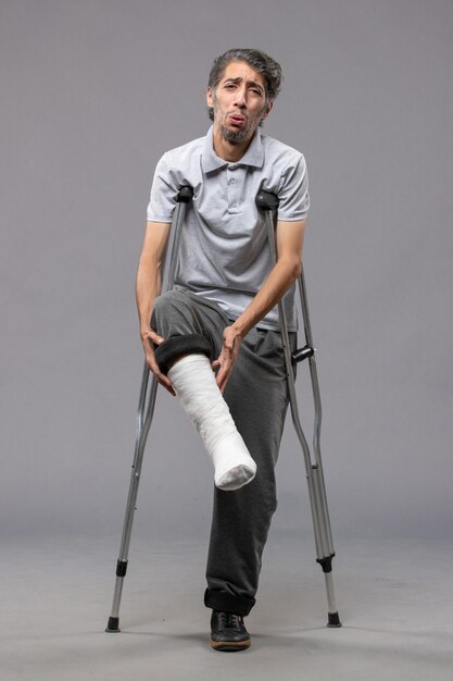 Вид спереди молодой человек, использующий костыли из-за сломанной ноги на сером столе, отключил ногу, сломал ногу, авария