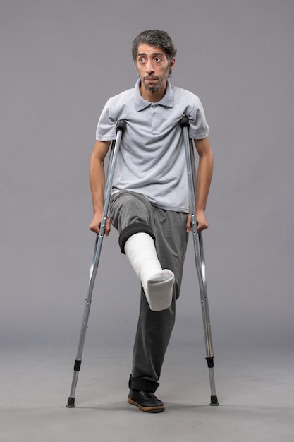 Front view young man using crutches due to broken foot on grey desk leg accident disable broke damage broken