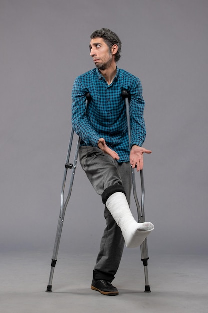 Free photo front view young man using crutches due to broken foot on grey desk leg accident disable broke damage