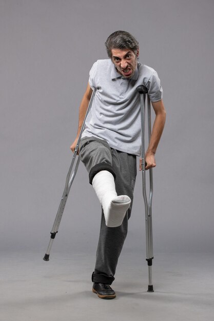 灰色の床の壊れた足のために松葉杖を使用している正面図の若い男は壊れた事故の足の足を無効にします