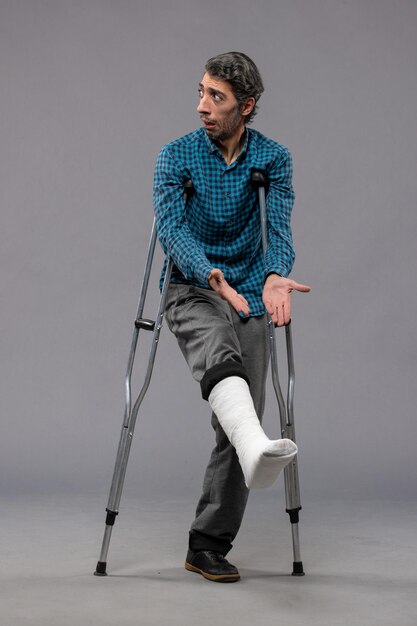 Front view young man using crutches due to broken foot on grey floor leg accident disable broke damage