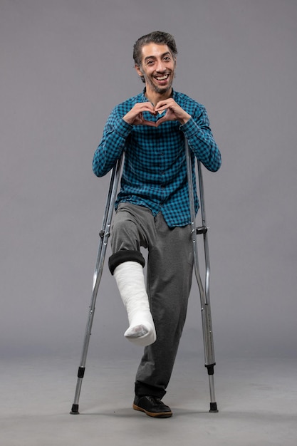 Front view young man using crutches due to broken foot on a grey wall accident disable broke damage leg