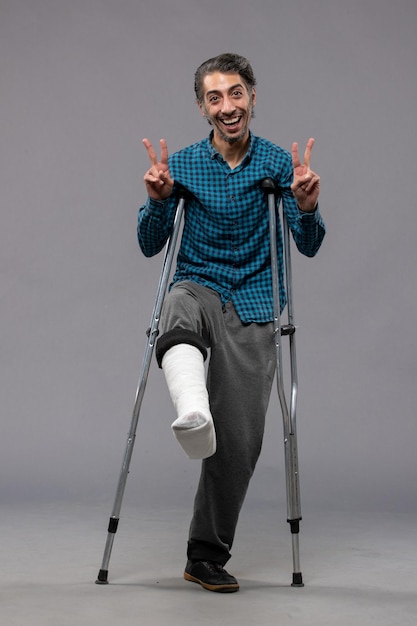 Free photo front view young man using crutches due to broken foot on grey wall accident disable broke damage leg