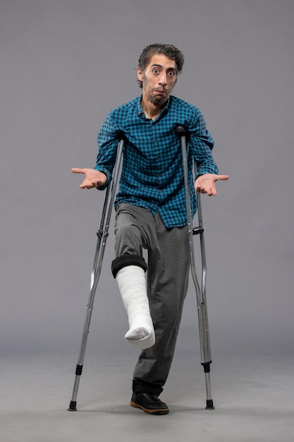 Free photo front view young man using crutches due to broken foot on grey wall accident disable broke damage legs