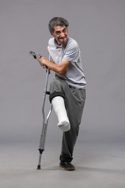 Front view young man using crutches due to broken foot on the grey wall accident disable broke leg broken