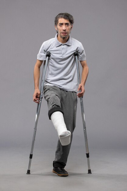 Front view young man using crutches due to broken foot on grey wall accident disable broke leg damage broken