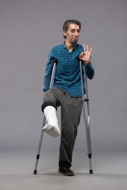 Вид спереди молодой человек, использующий костыли из-за сломанной ноги на серой стене, инвалид, сломал ногу