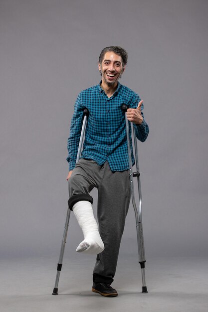 Front view young man using crutches due to broken foot on grey wall accident disabled broke damage leg