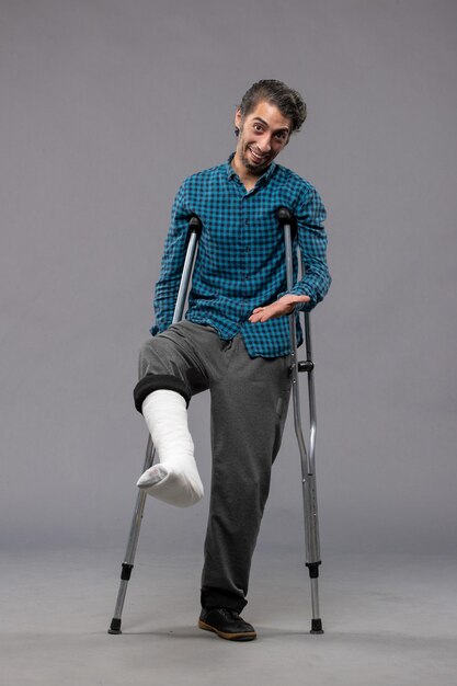 Front view young man using crutches due to broken foot on the grey wall accident disabled broke damage leg