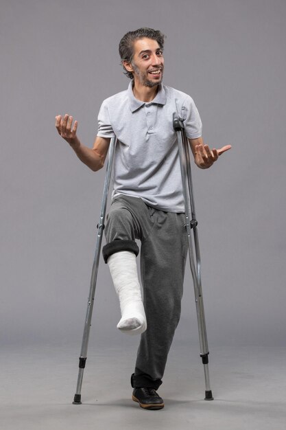 灰色の壁の事故で足の骨折のため松葉杖を使用している正面図の若い男は足の骨折を無効にします