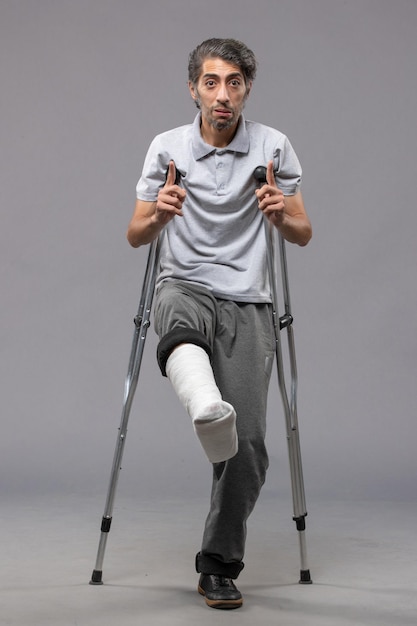 Front view young man using crutches due to broken foot on grey wall disable accident leg foot broken