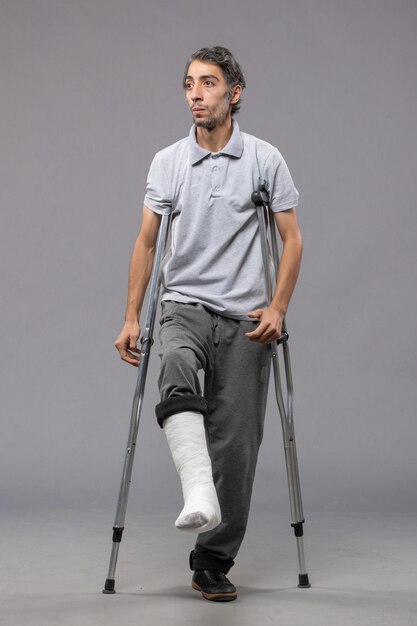 灰色の壁の壊れた足のために松葉杖を使用している正面図の若い男は壊れた事故の足の足を無効にします