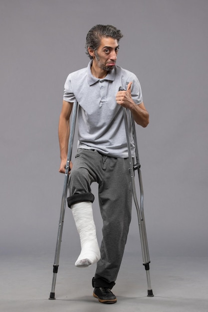 Front view young man using crutches due to broken foot on a grey wall disable broke leg broken accident