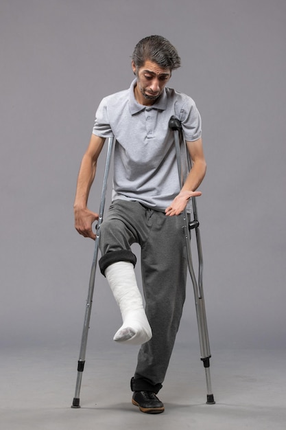 Free photo front view young man using crutches due to broken foot on the grey wall disable broke leg broken accident