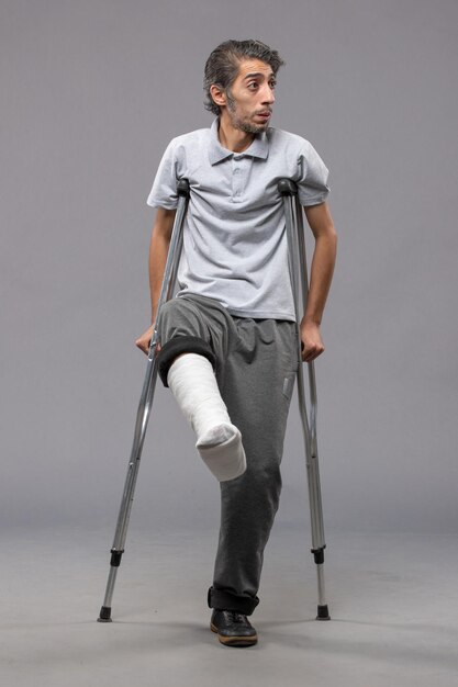 Front view young man using crutches due to broken foot on a grey wall disable broken accident leg foot