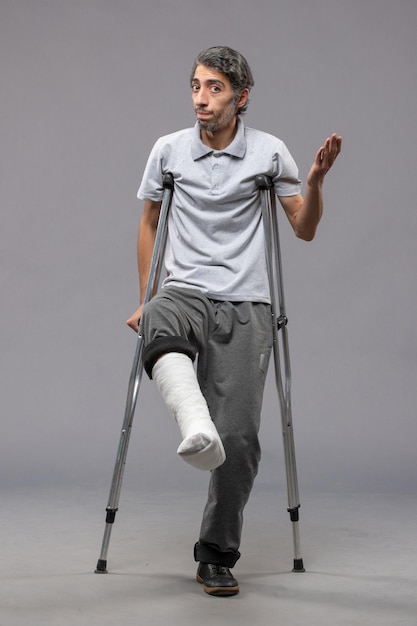 Вид спереди молодой человек, использующий костыли из-за сломанной ноги на серой стене, отключил сломанную ногу в результате аварии