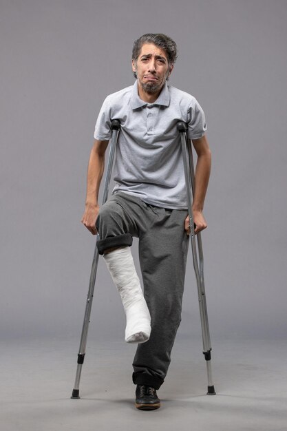 Вид спереди молодой человек, использующий костыли из-за сломанной ноги на серой стене, отключил ногу, сломал ногу, авария
