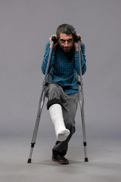 Front view young man using crutches due to broken foot on grey wall leg accident disable broke damage broken