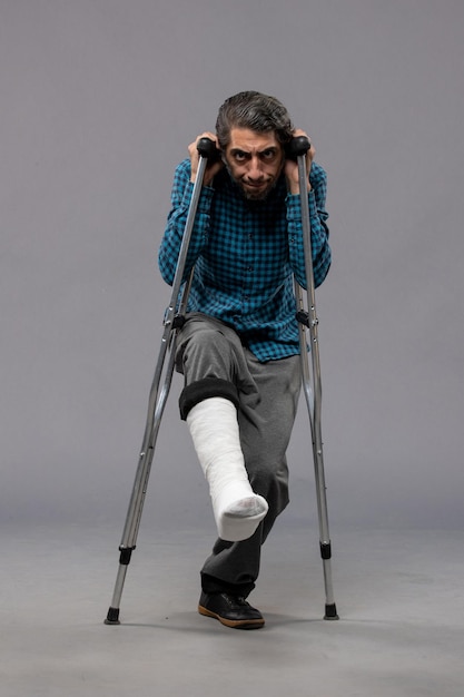 Вид спереди молодой человек, использующий костыли из-за сломанной ноги на серой стене, несчастный случай с ногой, сломанный, сломанный, сломанный