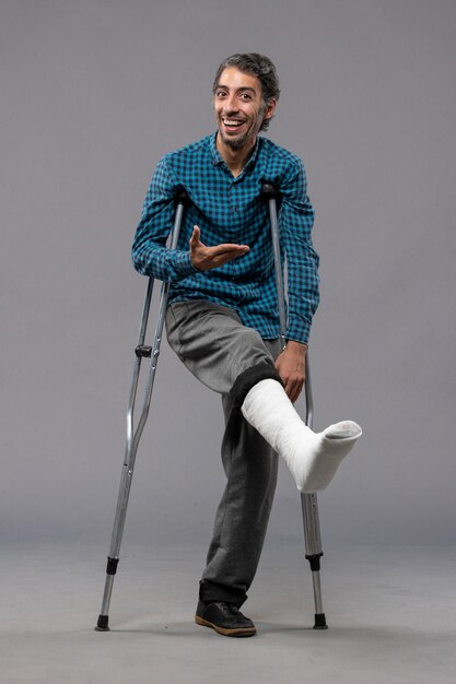 灰色の壁の脚の事故で足が折れたために松葉杖を使用している正面図の若い男は壊れた損傷を無効にします