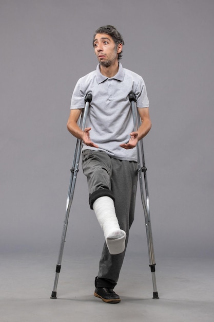 Front view young man using crutches due to broken foot on grey wall legs accident disable broke damage broken