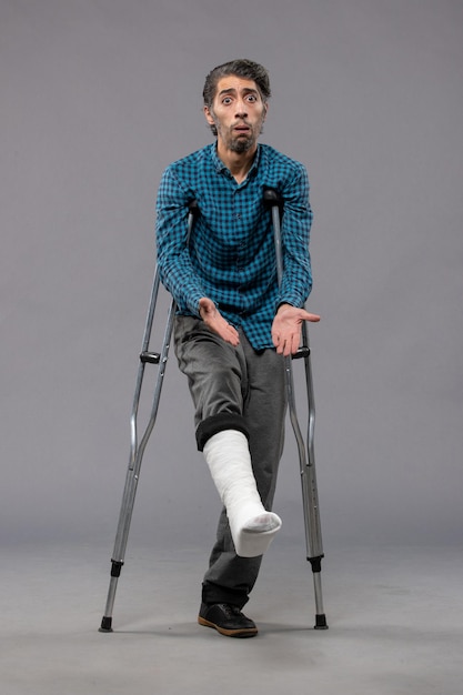 Front view young man using crutches due to broken foot on grey wall legs accident disable broke damage