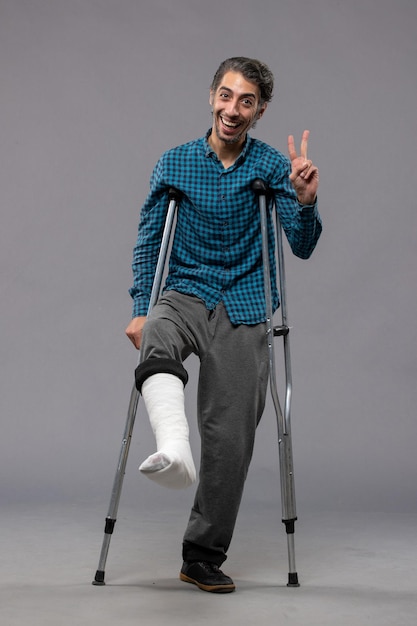 無料写真 灰色の壁の事故で笑っている足の骨折のために松葉杖を使用している正面図の若い男は、損傷した足を骨折しました