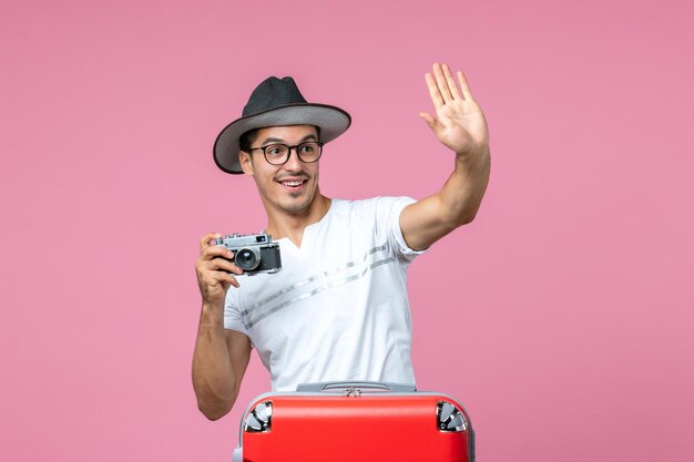 Front view of young man in vacation with bag holding camera taking photos on the pink wall