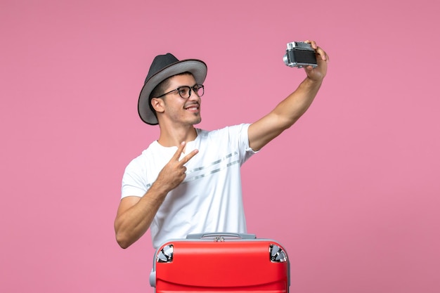 Front view of young man in vacation with red bag taking photos on pink wall