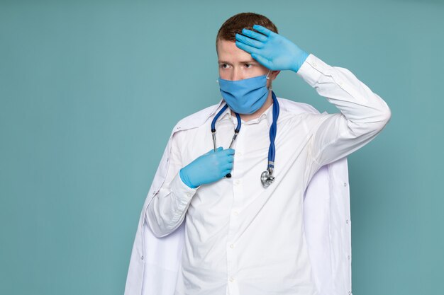 Вид спереди молодой человек в белом медицинском костюме синие перчатки и маска на синем полу