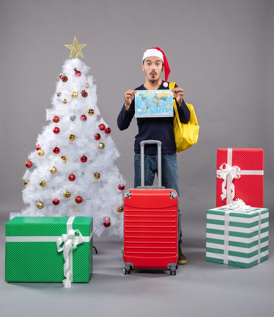 Вид спереди молодой человек с рюкзаком, держащий карту мира обеими руками возле белой рождественской елки на сером изолированном