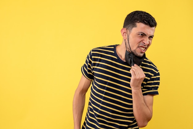 Front view young man with black and white striped t-shirt taking off his mask yellow background copy space