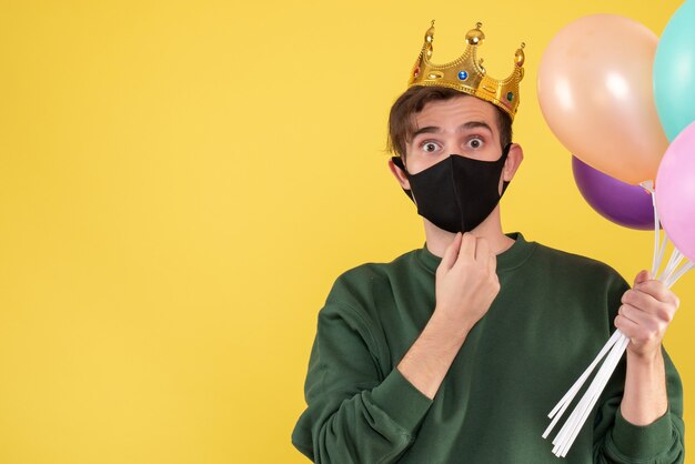 Вид спереди молодой человек с короной и черной маской, держащий воздушные шары на желтом