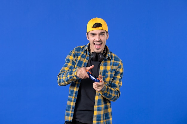 Front view young man with gamepad and earphones playing video game on blue wall