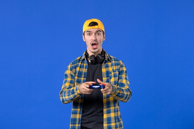 青い壁でビデオゲームをプレイするゲームパッドとイヤホンを持つ正面図の若い男
