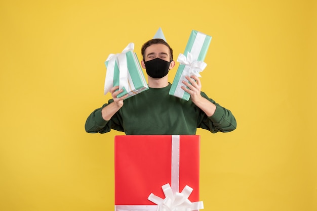 Front view young man with gift boxes standing behind big giftbox on yellow 