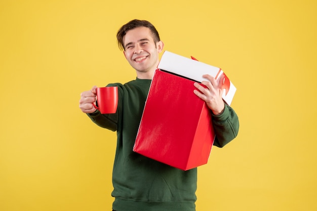 Front view young man with green sweater holding big gift and red cup standing on yellow 