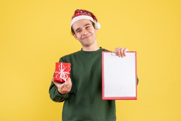 Front view young man with green sweater holding clipboard and gift standing on yellow 