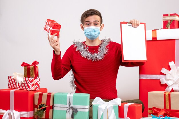 Front view young man with holding gift and clipboard back sitting around xmas gifts