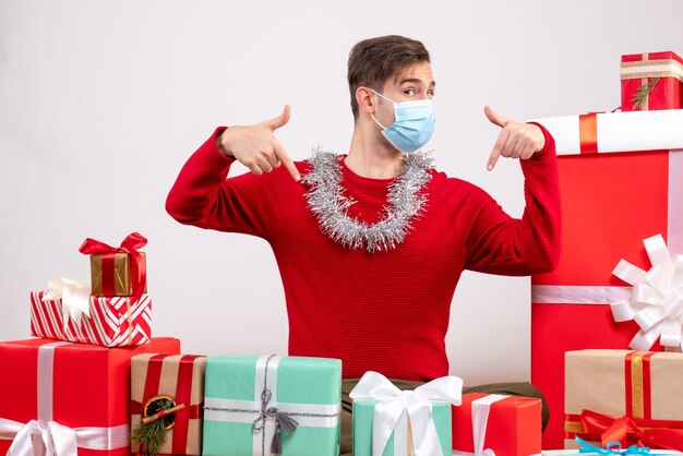 Giovane di vista frontale con il dito della maschera che indica se stesso seduto intorno ai regali di natale