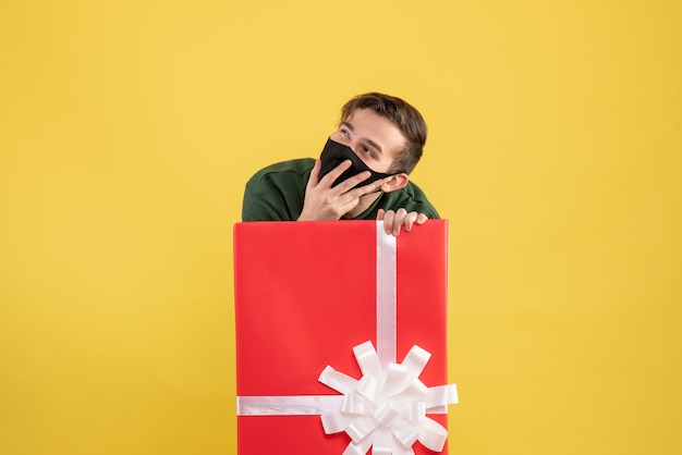 Front view young man with mask hiding behind big giftbox on yellow 