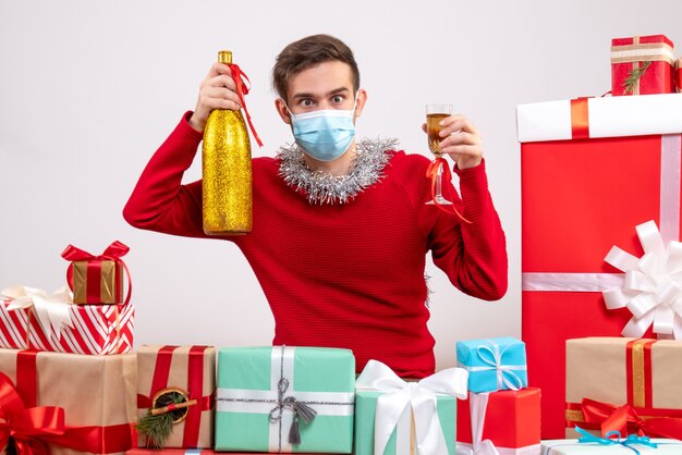 Front view young man with mask holding champagne sitting around xmas gifts