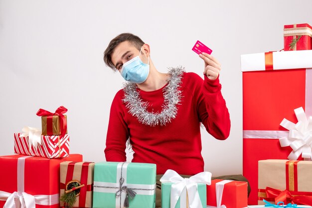 Вид спереди молодой человек с маской, держащий кредитную карту вокруг рождественских подарков