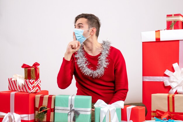 Вид спереди молодой человек с маской, делая знак тсс, сидя вокруг рождественских подарков на белом