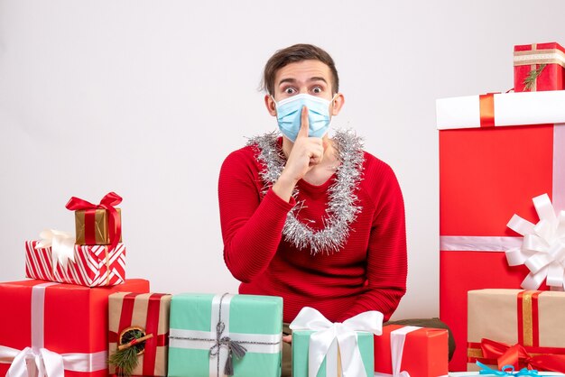 Giovane di vista frontale con la maschera che fa il segno di shh che si siede intorno ai regali di natale