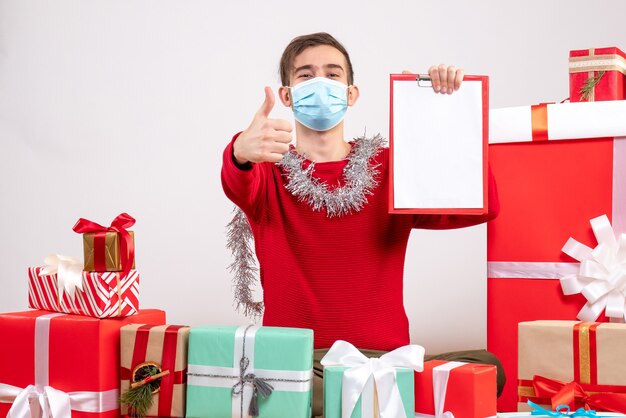 Front view young man with mask makng thumb up sign holding clipboard sitting around xmas gifts