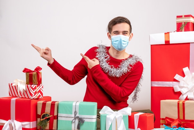 Front view young man with mask pointing at something sitting around xmas gifts