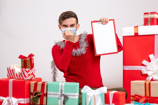 Front view young man with mask putting hand to his mouth sitting around xmas gifts