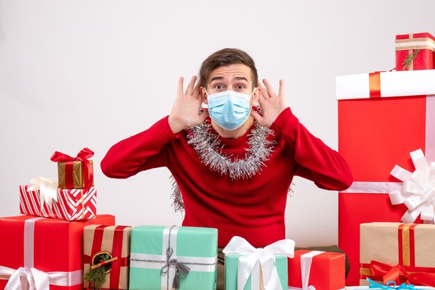 Front view young man with mask putting hands to ears listening something sitting around xmas gifts