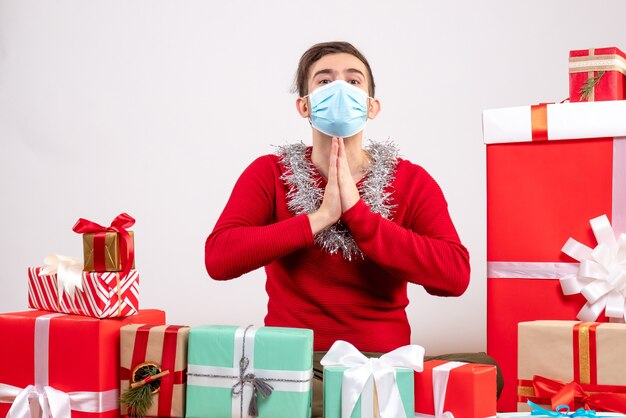 隔離された白い上のクリスマスプレゼントの周りに座っているマスクを持つ正面図若い男